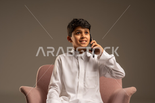 اتصالات ومكالمات هاتفية عبر الهاتف المحمول , الدردشة والتواصل مع الأهل والأصدقاء ، بورتريه لطفل عربي خليجي سعودي يرتدي الثوب التقليدي ويستخدم الجوال ،خلفية بيج .