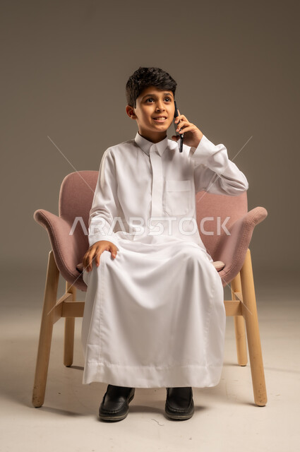 اتصالات ومكالمات هاتفية عبر الهاتف المحمول , الدردشة والتواصل مع الأهل والأصدقاء ، بورتريه لطفل عربي خليجي سعودي يرتدي الثوب التقليدي ويستخدم الجوال ،خلفية بيج .