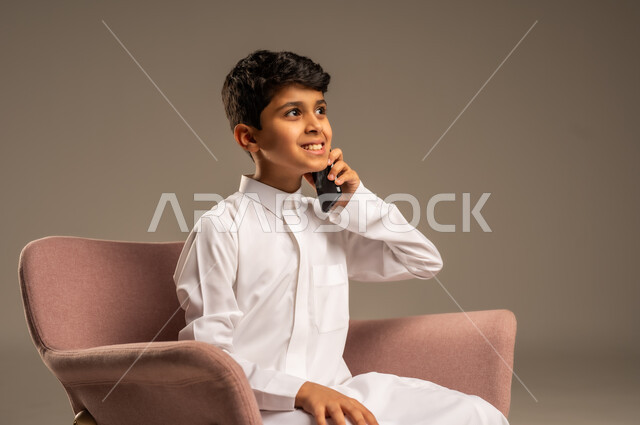 اتصالات ومكالمات هاتفية عبر الهاتف المحمول , الدردشة والتواصل مع الأهل والأصدقاء ، بورتريه لطفل عربي خليجي سعودي يرتدي الثوب التقليدي ويستخدم الجوال ،خلفية بيج .