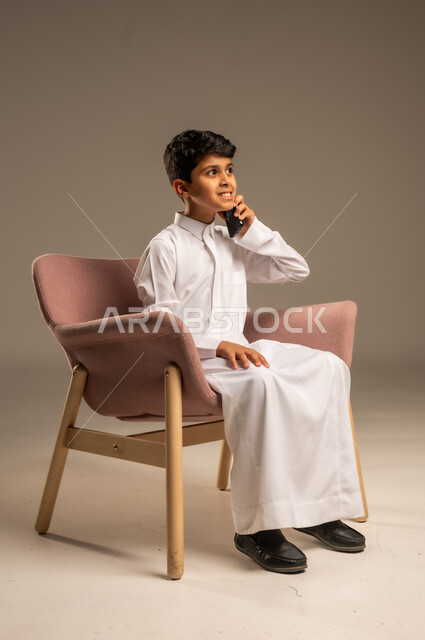 اتصالات ومكالمات هاتفية عبر الهاتف المحمول , الدردشة والتواصل مع الأهل والأصدقاء ، بورتريه لطفل عربي خليجي سعودي يرتدي الثوب التقليدي ويستخدم الجوال ،خلفية بيج .