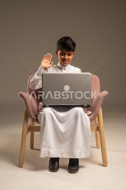 متابعة الدروس والشروحات عن بعد, بورتريه لطفل عربي خليجي سعودي يرتدي الثوب التقليدي يجلس علىالكرسي  ويستخدم اللابتوب, استعمال الأجهزة التقنية في التعلم, التسلية والترفيه بقضاء الوقت بتصفح مواقع التواصل الاجتماعي على الحاسوب المحمول, خلفية بيج