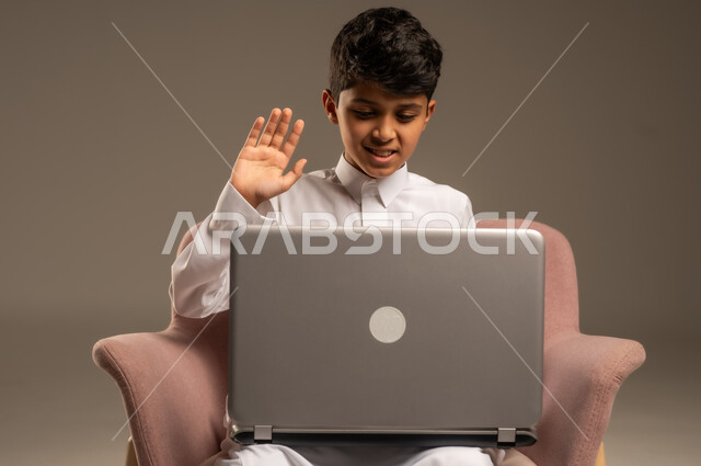 متابعة الدروس والشروحات عن بعد, بورتريه لطفل عربي خليجي سعودي يرتدي الثوب التقليدي يجلس على الكرسي ويستخدم اللابتوب, استعمال الأجهزة التقنية في التعلم, التسلية والترفيه بقضاء الوقت بتصفح مواقع التواصل الاجتماعي على الحاسوب المحمول, خلفية بيج