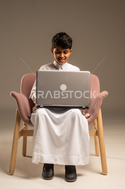 متابعة الدروس والشروحات عن بعد, بورتريه لطفل عربي خليجي سعودي يرتدي الثوب التقليدي يجلس على الكرسي ويستخدم اللابتوب, استعمال الأجهزة التقنية في التعلم, التسلية والترفيه بقضاء الوقت بتصفح مواقع التواصل الاجتماعي على الحاسوب المحمول, خلفية بيج