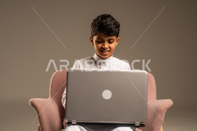 Following lessons and explanations remotely, portrait of a Saudi Arabian Gulf child wearing traditional dress sitting on a chair and using a laptop, using technical devices in learning, entertainment and fun by spending time browsing social media sites on a laptop, beige background