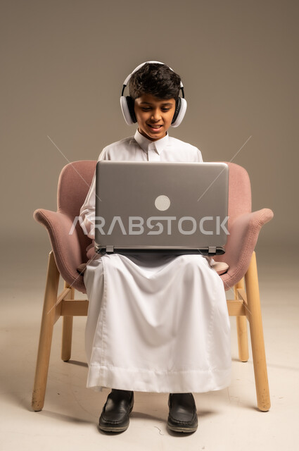 متابعة الدروس والشروحات عن بعد, بورتريه لطفل عربي خليجي سعودي يرتدي الثوب التقليدي يجلس على الكرسي ويستخدم اللابتوب, استعمال الأجهزة التقنية في التعلم, التسلية والترفيه بقضاء الوقت بتصفح مواقع التواصل الاجتماعي على الحاسوب المحمول, خلفية بيج