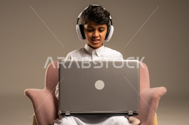 متابعة الدروس والشروحات عن بعد, بورتريه لطفل عربي خليجي سعودي يرتدي الثوب التقليدي يجلس على الكرسي ويستخدم اللابتوب, استعمال الأجهزة التقنية في التعلم, التسلية والترفيه بقضاء الوقت بتصفح مواقع التواصل الاجتماعي على الحاسوب المحمول, خلفية بيج