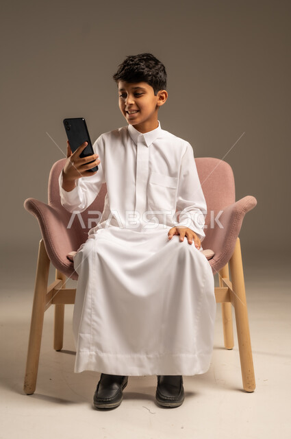 Using modern electronic devices in learning, integrating science with technology, following up on assignments online via mobile, portrait of a Saudi Arabian Gulf boy wearing traditional dress holding a mobile phone in his hand, beige background