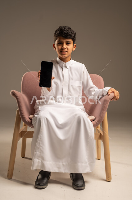 استعمال الأجهزة الإلكترونية الحديثة بالتعلم، دمج العلم بالتقنية والتكنولوجيا، متابعة الواجبات اونلاين عبر الجوال، بورتريه لولد عربي خليجي سعودي  يرتدي الثوب التقليدي يمسك هاتف محمول بيده، خلفية بيج