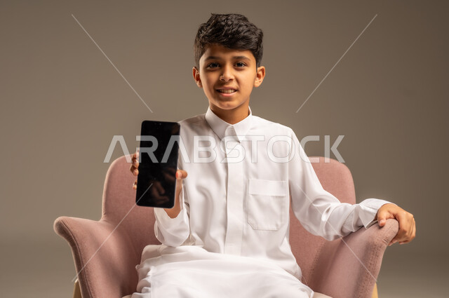 استعمال الأجهزة الإلكترونية الحديثة بالتعلم، دمج العلم بالتقنية والتكنولوجيا، متابعة الواجبات اونلاين عبر الجوال، بورتريه لولد عربي خليجي سعودي  يرتدي الثوب التقليدي يمسك هاتف محمول بيده، خلفية بيج