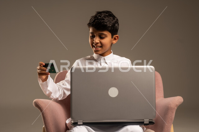 الشراء والدفع من خلال تطبيقات الجوال، التقنيات الحديثة والمتطورة، بورتريه لطفل عربي خليجي سعودي  يرتدي الثوب التقليدي يجلس على كرسي يمسك بيده اللابتوب وبطاقة الإئتمان البنكية، تسديد الفواتير بالبطاقات الإلكترونية، خلفية بيج