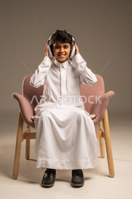 استخدام تقنية الهيدفون، الاستماع الى الأغاني والموسيقى عبر سماعة الرأس، بورتريه مقرب لطفل عربي خليجي سعودي يضع سماعة الرأس يقوم بالإصغاء والاستماع إلى شيء ما، خلفية بيج