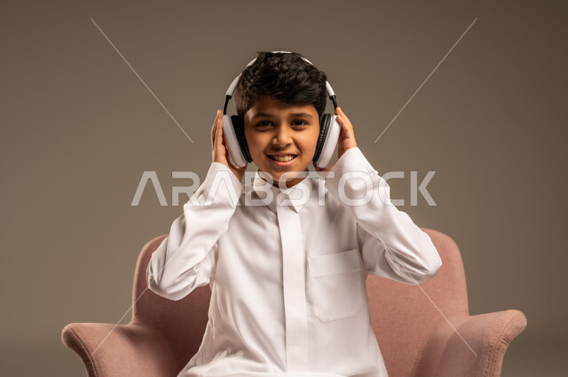 استخدام تقنية الهيدفون، الاستماع الى الأغاني والموسيقى عبر سماعة الرأس، بورتريه مقرب لطفل عربي خليجي سعودي يضع سماعة الرأس يقوم بالإصغاء والاستماع إلى شيء ما، خلفية بيج