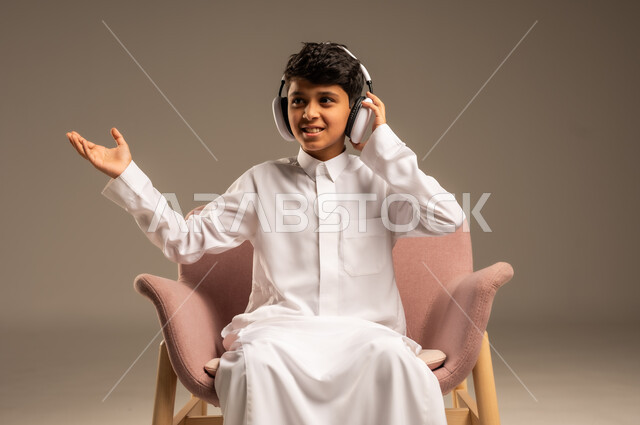 استخدام تقنية الهيدفون، الاستماع الى الأغاني والموسيقى عبر سماعة الرأس، بورتريه مقرب لطفل عربي خليجي سعودي يضع سماعة الرأس يقوم بالإصغاء والاستماع إلى شيء ما، خلفية بيج