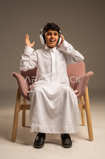 استخدام تقنية الهيدفون، الاستماع الى الأغاني والموسيقى عبر سماعة الرأس، بورتريه مقرب لطفل عربي خليجي سعودي يضع سماعة الرأس يقوم بالإصغاء والاستماع إلى شيء ما، خلفية بيج