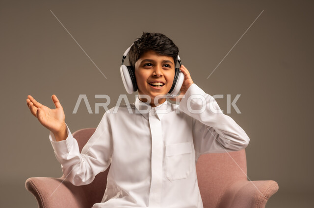 استخدام تقنية الهيدفون، الاستماع الى الأغاني والموسيقى عبر سماعة الرأس، بورتريه مقرب لطفل عربي خليجي سعودي يضع سماعة الرأس يقوم بالإصغاء والاستماع إلى شيء ما، خلفية بيج
