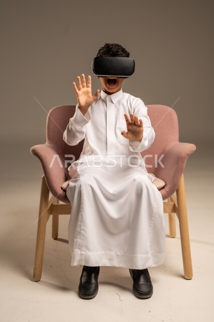 Gestures of interaction and integration, development of teaching methods using modern technologies and simulation of the imaginary world, portrait of a Saudi Arabian Gulf boy wearing traditional dress wearing 3D virtual reality glasses, beige background