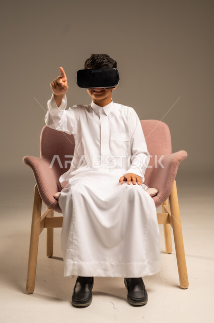 إيماءات التفاعل والاندماج، تطورأساليب التعليم باستخدام التقنيات الحديثة ومحاكاة العالم الخيالي، بورتريه لولد عربي خليجي سعودي يرتدي الثوب التقليدي يلبس نظارة الواقع الإفتراضي ثلاثية الأبعاد, خلفية بيج