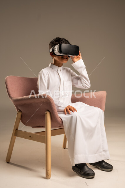 إيماءات التفاعل والاندماج، تطورأساليب التعليم باستخدام التقنيات الحديثة ومحاكاة العالم الخيالي، بورتريه لولد عربي خليجي سعودي يرتدي الثوب التقليدي يلبس نظارة الواقع الإفتراضي ثلاثية الأبعاد, خلفية بيج