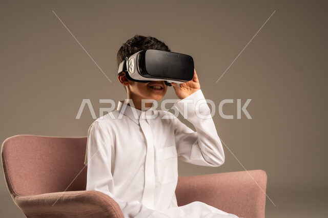 Gestures of interaction and integration, development of teaching methods using modern technologies and simulation of the imaginary world, portrait of a Saudi Arabian Gulf boy wearing traditional dress wearing 3D virtual reality glasses, beige background