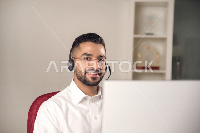 موظف عربي خليجي سعودي يعمل في خدمة العملاء ويرتدي سماعة الاتصال، استعلامات واستفسارات، الاجابه على اسئلة العملاء، اجراء اتصال فيديو من خلال الحاسوب، نظارة طبية، متابعة وتطوير العمل