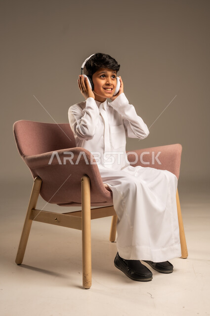 استخدام تقنية الهيدفون، الاستماع الى الأغاني والموسيقى عبر سماعة الرأس، بورتريه مقرب لطفل عربي خليجي سعودي يضع سماعة الرأس يقوم بالإصغاء والاستماع إلى شيء ما، خلفية بيج