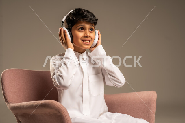 استخدام تقنية الهيدفون، الاستماع الى الأغاني والموسيقى عبر سماعة الرأس، بورتريه مقرب لطفل عربي خليجي سعودي يضع سماعة الرأس يقوم بالإصغاء والاستماع إلى شيء ما، خلفية بيج