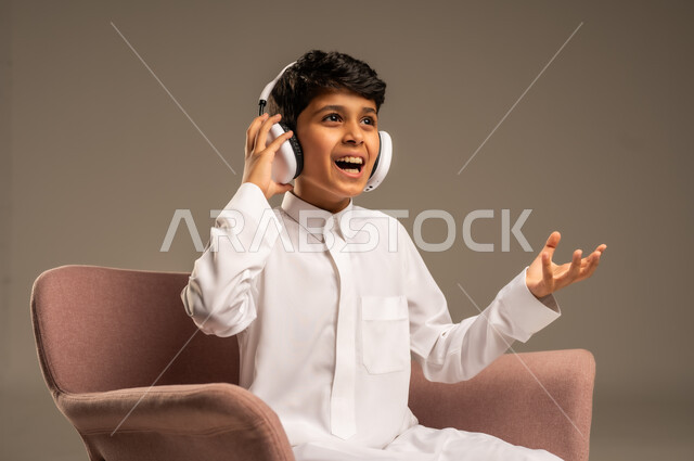 استخدام تقنية الهيدفون، الاستماع الى الأغاني والموسيقى عبر سماعة الرأس، بورتريه مقرب لطفل عربي خليجي سعودي يضع سماعة الرأس يقوم بالإصغاء والاستماع إلى شيء ما، خلفية بيج