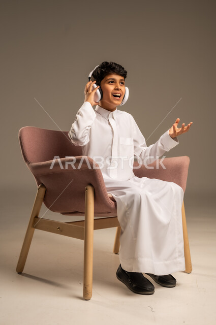 استخدام تقنية الهيدفون، الاستماع الى الأغاني والموسيقى عبر سماعة الرأس، بورتريه مقرب لطفل عربي خليجي سعودي يضع سماعة الرأس يقوم بالإصغاء والاستماع إلى شيء ما، خلفية بيج