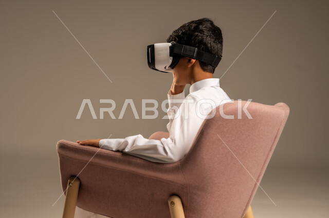 Gestures of interaction and integration, development of teaching methods using modern technologies and simulation of the imaginary world, portrait of a Saudi Arabian Gulf boy wearing traditional dress wearing 3D virtual reality glasses, beige background