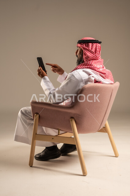 اتصالات ومكالمات هاتفية عبر الهاتف المحمول , الدردشة والتواصل مع الأهل والأصدقاء ، بورتريه لرجل عربي خليجي سعودي يرتدي الثوب التقليدي والشماغ ويستخدم الجوال، خلفية بيج