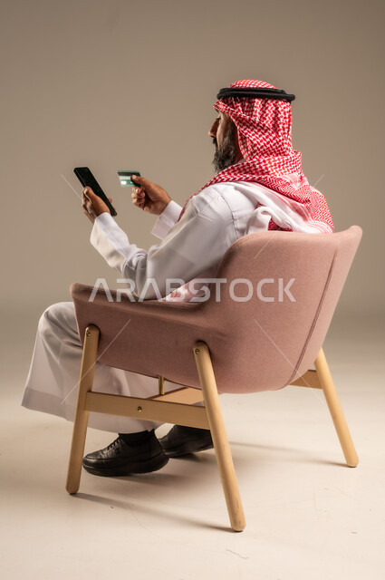 Purchase and payment through mobile programs and applications, modern and advanced technologies, portrait of a Saudi Arabian Gulf man wearing a traditional thobe and shemagh sitting on a chair holding a mobile phone and a bank credit card, paying bills with electronic cards, beige background