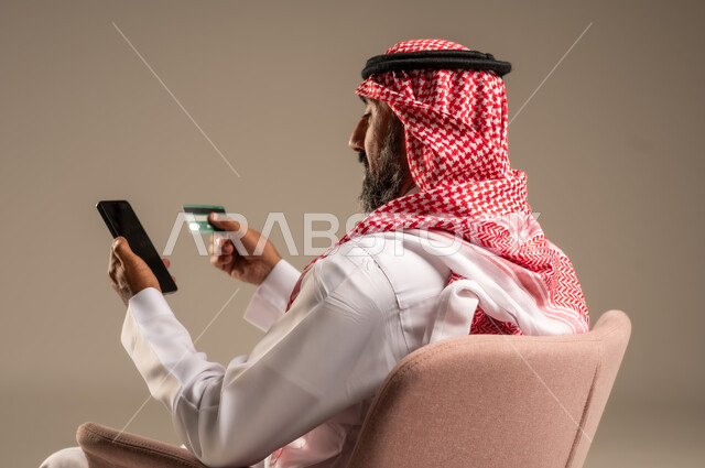 Purchase and payment through mobile programs and applications, modern and advanced technologies, portrait of a Saudi Arabian Gulf man wearing a traditional thobe and shemagh sitting on a chair holding a mobile phone and a bank credit card, paying bills with electronic cards, beige background