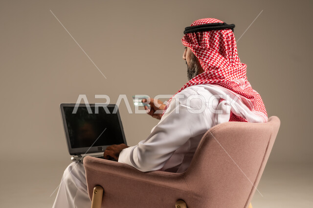 عقد إجتماع عمل عبر الإنترنت، تصفح برامج التواصل الاجتماعي, إستخدام الحاسوب المحمول لإنجاز المهام، بورتريه لرجل عربي خليجي سعودي يرتدي الثوب التقليدي والشماغ يحمل جهاز اللابتوب يجلس على كرسي مريح، تطور التكنولوجيا والأجهزة التقنية، خلفية بيج