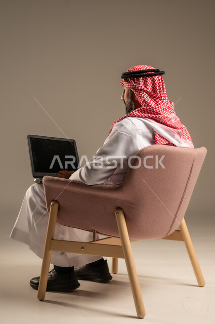 عقد إجتماع عمل عبر الإنترنت، تصفح برامج التواصل الاجتماعي, إستخدام الحاسوب المحمول لإنجاز المهام، بورتريه لرجل عربي خليجي سعودي يرتدي الثوب التقليدي والشماغ يحمل جهاز اللابتوب يجلس على كرسي مريح، تطور التكنولوجيا والأجهزة التقنية، خلفية بيج
