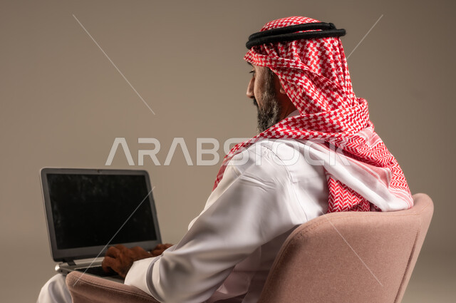 عقد إجتماع عمل عبر الإنترنت، تصفح برامج التواصل الاجتماعي, إستخدام الحاسوب المحمول لإنجاز المهام، بورتريه لرجل عربي خليجي سعودي يرتدي الثوب التقليدي والشماغ يحمل جهاز اللابتوب يجلس على كرسي مريح، تطور التكنولوجيا والأجهزة التقنية، خلفية بيج