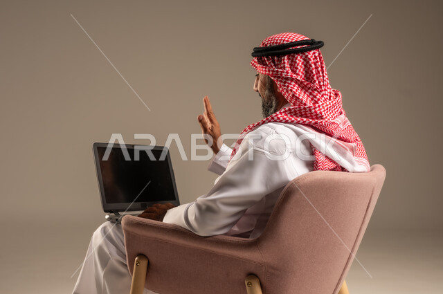 عقد إجتماع عمل عبر الإنترنت، تصفح برامج التواصل الاجتماعي, إستخدام الحاسوب المحمول لإنجاز المهام، بورتريه لرجل عربي خليجي سعودي يرتدي الثوب التقليدي والشماغ يحمل جهاز اللابتوب يجلس على كرسي مريح، تطور التكنولوجيا والأجهزة التقنية، خلفية بيج