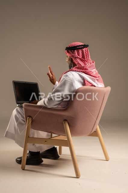 Holding a business meeting online, browsing social media, using laptop to complete tasks, portrait of a Saudi Arabian Gulf man wearing traditional thobe and shemagh holding a laptop sitting on a comfortable chair, development of technology and technical devices, beige background