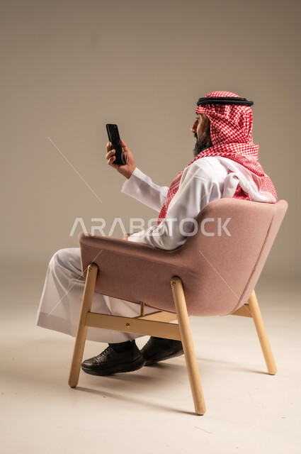 Mobile phone calls and communications, chatting and communicating with family and friends, portrait of a Saudi Arabian Gulf man wearing traditional thobe and shemagh and using a mobile phone, beige background