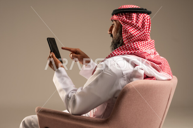 Mobile phone calls and communications, chatting and communicating with family and friends, portrait of a Saudi Arabian Gulf man wearing traditional thobe and shemagh and using a mobile phone, beige background