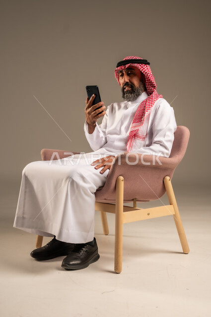 Using modern applications and electronic marketing, portrait of an elderly Saudi Arabian Gulf man wearing traditional thobe and shemagh using a mobile phone, social networking programs via mobile phone, beige background.