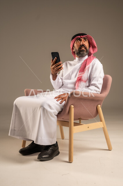 استخدام التطبيقات الحديثة و التسويق الألكتروني, بورتريه لرجل عربي سعودي خليجي كبير بالسن  يرتدي الثوب التقليدي والشماغ يستخدم الجوال, برامج التواصل الاجتماعي عبر الهاتف المحمول, خلفية بيج .
