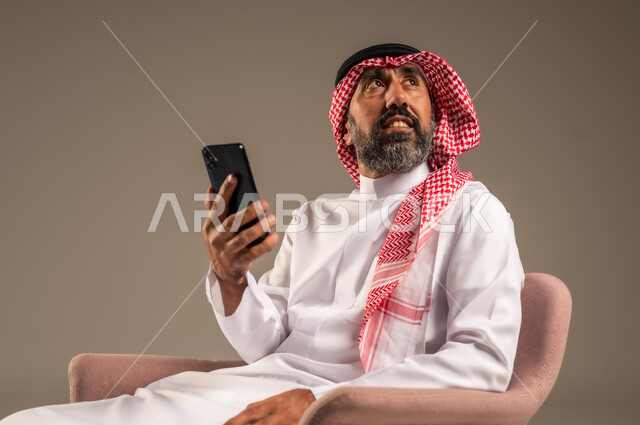 استخدام التطبيقات الحديثة و التسويق الألكتروني, بورتريه لرجل عربي سعودي خليجي كبير بالسن  يرتدي الثوب التقليدي والشماغ يستخدم الجوال, برامج التواصل الاجتماعي عبر الهاتف المحمول, خلفية بيج .