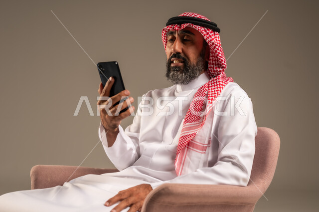 استخدام التطبيقات الحديثة و التسويق الألكتروني, بورتريه لرجل عربي سعودي خليجي كبير بالسن  يرتدي الثوب التقليدي والشماغ يستخدم الجوال, برامج التواصل الاجتماعي عبر الهاتف المحمول, خلفية بيج .