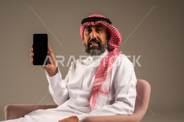 استخدام التطبيقات الحديثة و التسويق الألكتروني, بورتريه لرجل عربي سعودي خليجي كبير بالسن  يرتدي الثوب التقليدي والشماغ يمسك الجوال بشاشة سوداء فارغة, برامج التواصل الاجتماعي عبر الهاتف المحمول, خلفية بيج .