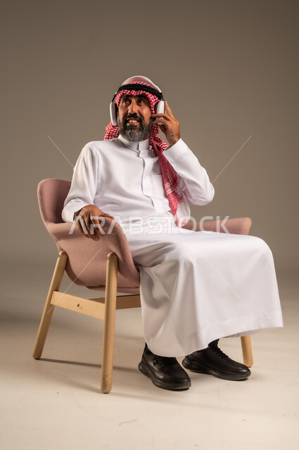 قضاء أوقات ممتعة ومسلية، الإصغاء والاستمتاع باستخدام الهيدفون، الاستماع إلى المواهب الغنائية الفنية، بورتريه لرجل عربي خليجي سعودي كبير بالسن يرتدي الثوب التقليدي والشماغ  يضع سماعة الرأس ويستمتع بسماع اغاني منوعة، خلفية بيج