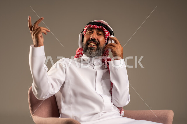 قضاء أوقات ممتعة ومسلية، الإصغاء والاستمتاع باستخدام الهيدفون، الاستماع إلى المواهب الغنائية الفنية، بورتريه لرجل عربي خليجي سعودي كبير بالسن يرتدي الثوب التقليدي والشماغ  يضع سماعة الرأس ويستمتع بسماع اغاني منوعة، خلفية بيج