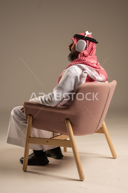 Having a fun and entertaining time, listening and enjoying using headphones, listening to artistic singing talents, portrait of an elderly Saudi Arabian Gulf man wearing traditional thobe and shemagh, putting on headphones and enjoying listening to various songs, beige background
