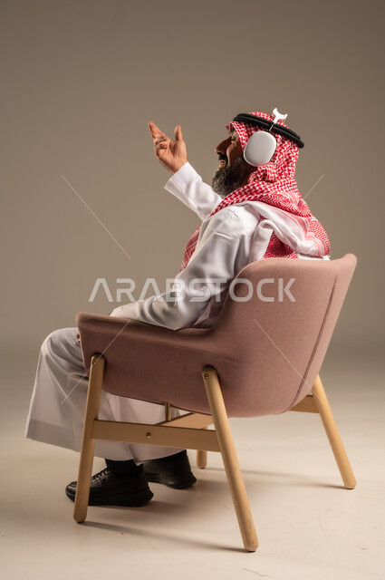 قضاء أوقات ممتعة ومسلية، الإصغاء والاستمتاع باستخدام الهيدفون، الاستماع إلى المواهب الغنائية الفنية، بورتريه لرجل عربي خليجي سعودي كبير بالسن يرتدي الثوب التقليدي والشماغ  يضع سماعة الرأس ويستمتع بسماع اغاني منوعة، خلفية بيج