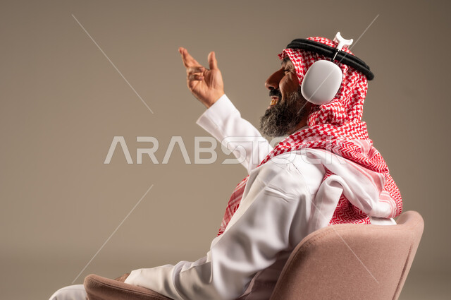 Having a fun and entertaining time, listening and enjoying using headphones, listening to artistic singing talents, portrait of an elderly Saudi Arabian Gulf man wearing traditional thobe and shemagh, putting on headphones and enjoying listening to various songs, beige background