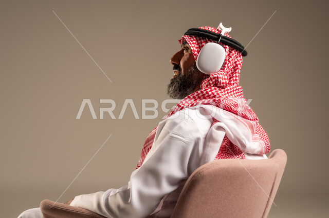 Having a fun and entertaining time, listening and enjoying using headphones, listening to artistic singing talents, portrait of an elderly Saudi Arabian Gulf man wearing traditional thobe and shemagh, putting on headphones and enjoying listening to various songs, beige background